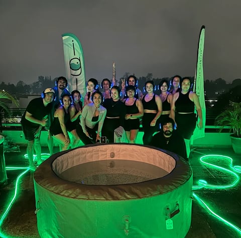 Group of people in neon green athletic wear posing behind a large green inflatable hot tub at night with green neon lighting